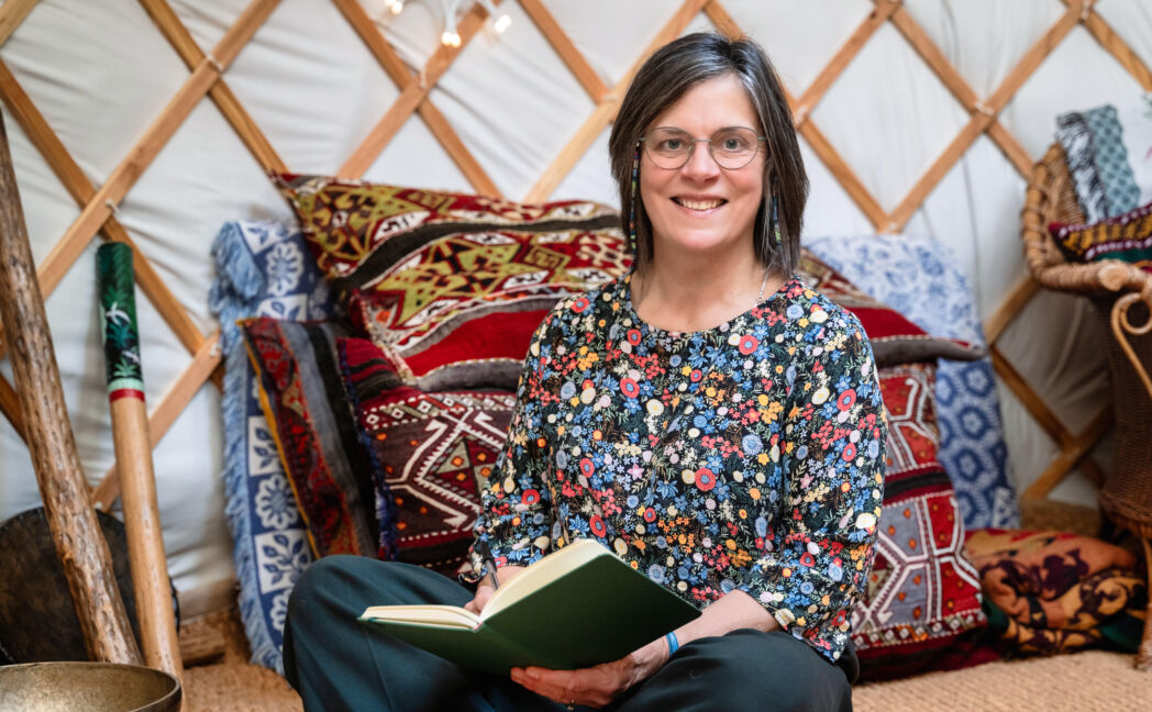 Woman sitting with a notebook inside a warmly decorated yurt, surrounded by colourful cushions and natural textures, creating a calm and inviting space.