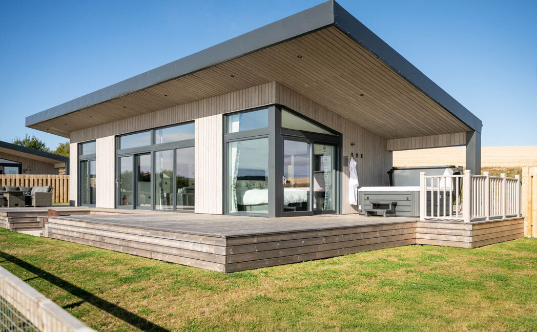 Exterior view of a modern luxury lodge with floor-to-ceiling windows, spacious decking, enclosed garden and private hot tub overlooking Aberdeenshire countryside.