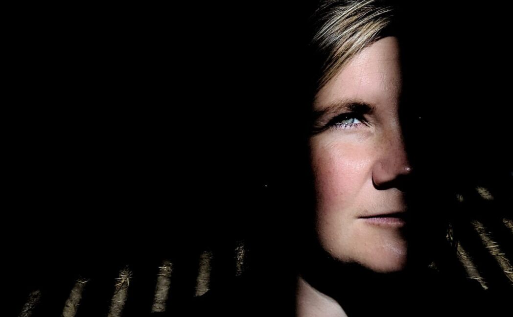 A dramatic portrait of a woman partially illuminated by sunlight filtering through slatted shadows. Half of her face is highlighted, while the other half fades into darkness, creating a striking contrast.