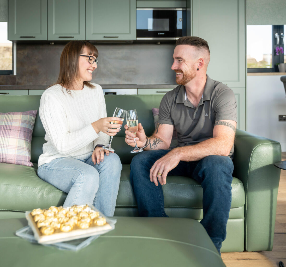 A couple clinks their glasses while sitting on a green couch in a modern living room, with a tray of chocolates on the table and a bottle of champagne nearby.