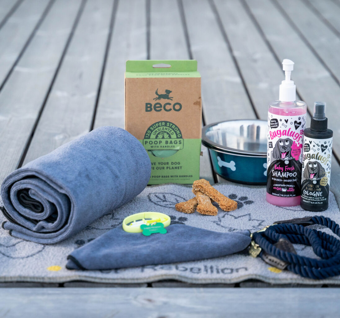 A collection of pet care items arranged on a wooden surface, including a towel, poo bags, bowl, dog shampoo, conditioner, treats, and lead.