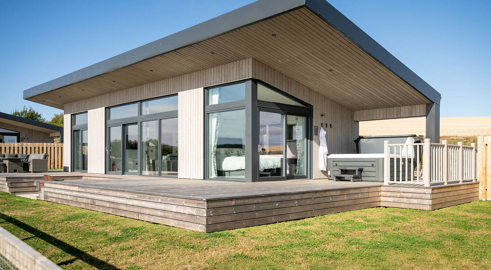 Exterior view of a modern luxury lodge with floor-to-ceiling windows, spacious decking, enclosed garden and private hot tub overlooking Aberdeenshire countryside.