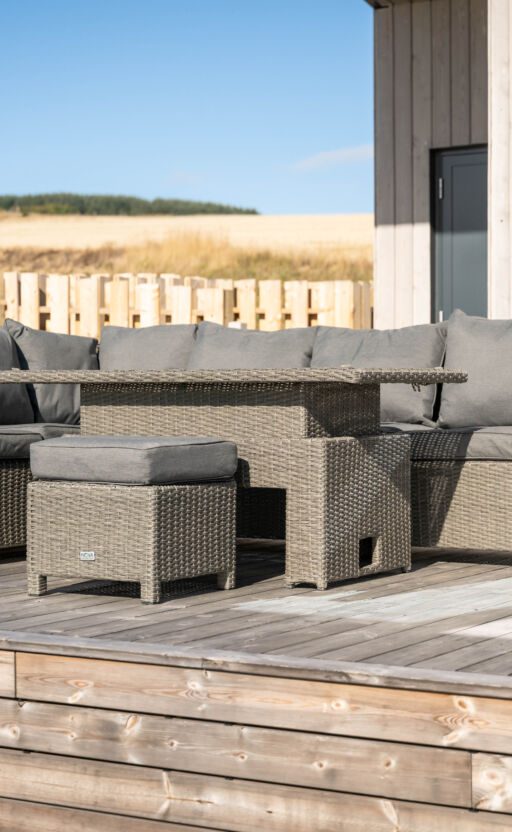 A patio set made of wicker, including a table and several chairs, arranged on a wooden deck outside a modern building, surrounded by greenery and a wooden fence.