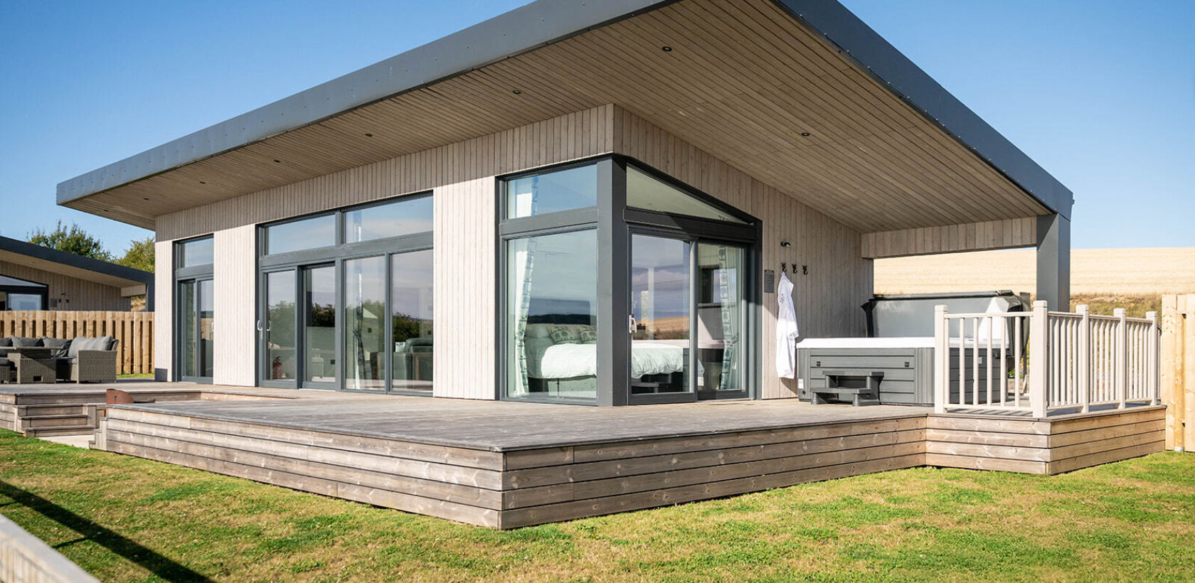 Exterior view of a modern luxury lodge with floor-to-ceiling windows, spacious decking, enclosed garden and private hot tub overlooking Aberdeenshire countryside.