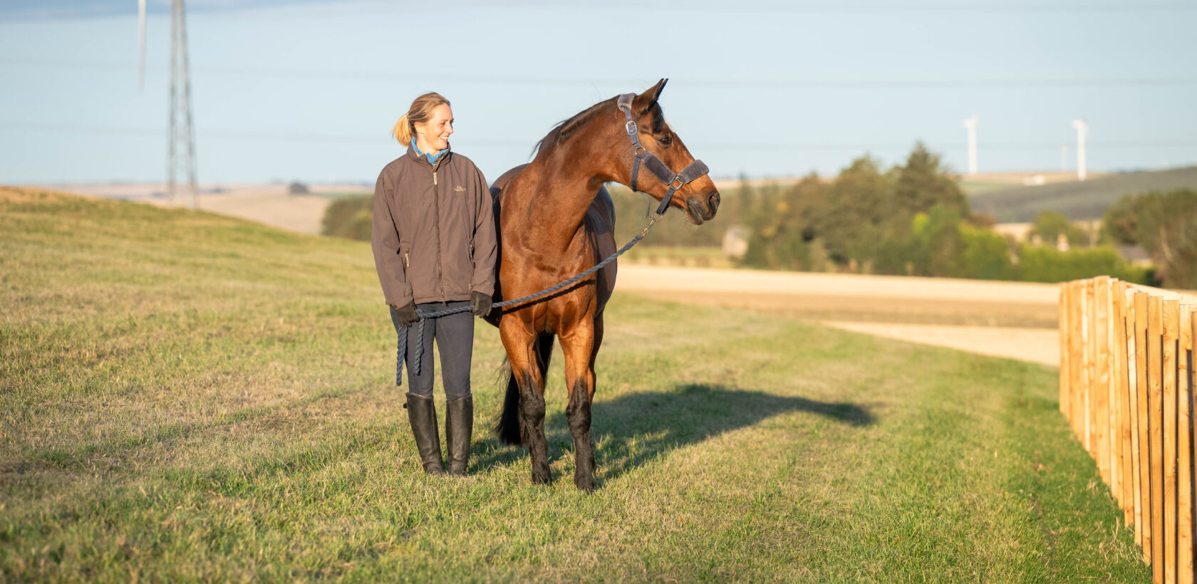 A person standing beside a horse on a grassy hill, with wind turbines visible in the background.
