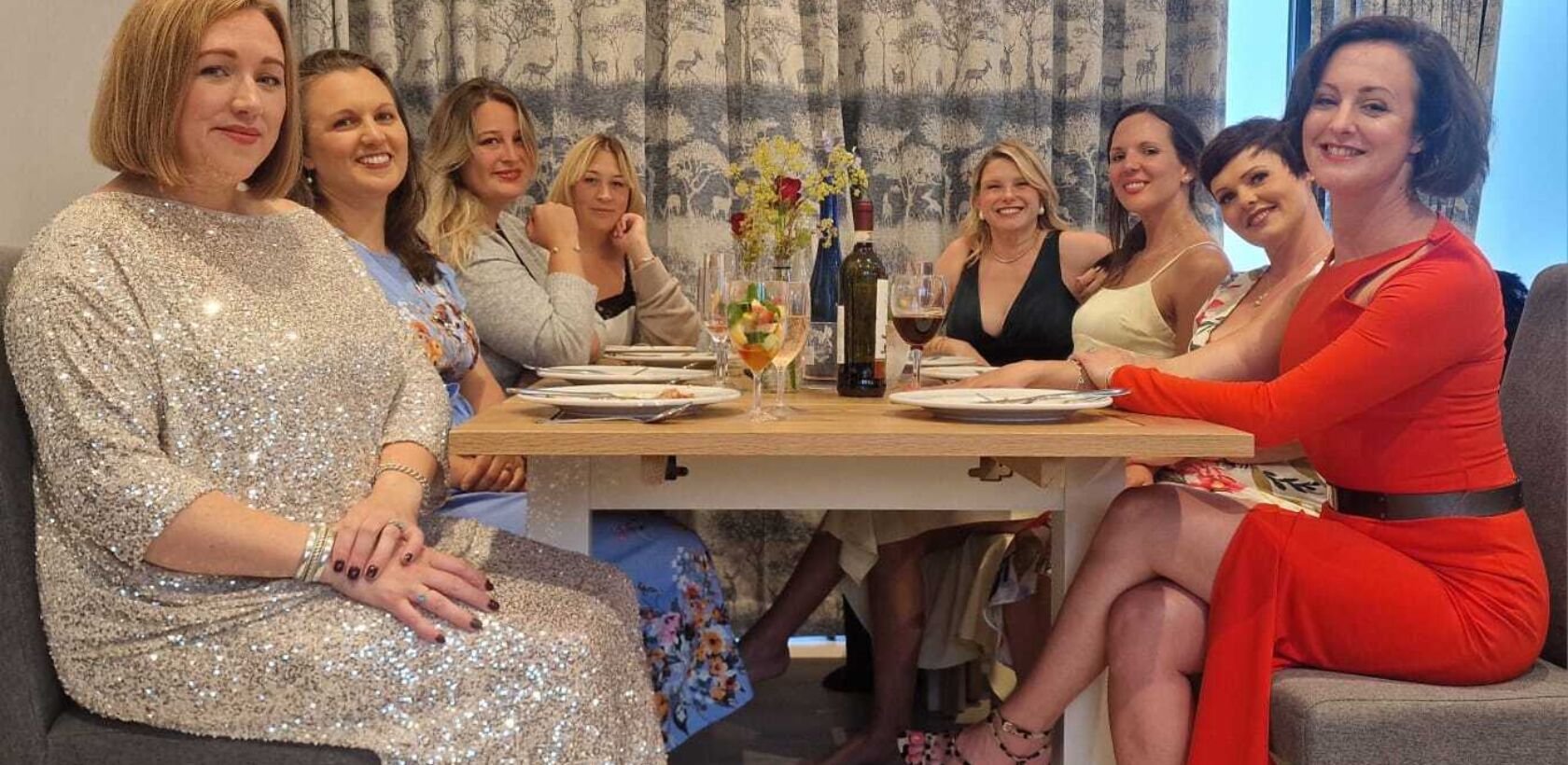 A group of women dressed in elegant outfits sitting around a dining table, enjoying drinks and food.